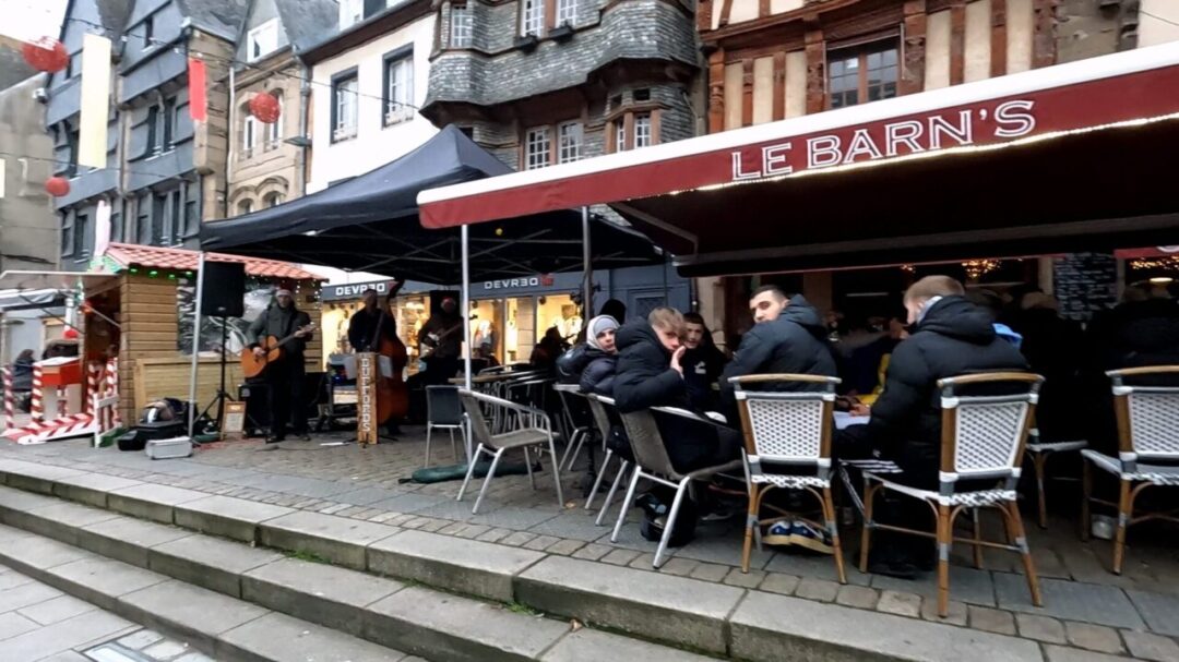A family waving at the camera in Place du Centre Christmas Market, with a small band playing on the left. Place du Centre 크리스마스 마켓에서 카메라를 보고 손을 흔드는 가족, 왼쪽에서는 작은 밴드가 연주 중.