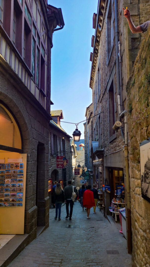 A narrow alley inside Mont Saint-Michel, lined with historic stone buildings, charming shops, and vintage lanterns hanging from chains above. 몽생미셸 내부의 좁은 골목길, 고풍스러운 석조 건물들과 아기자기한 상점들, 그리고 머리 위로 걸려 있는 빈티지 조명.