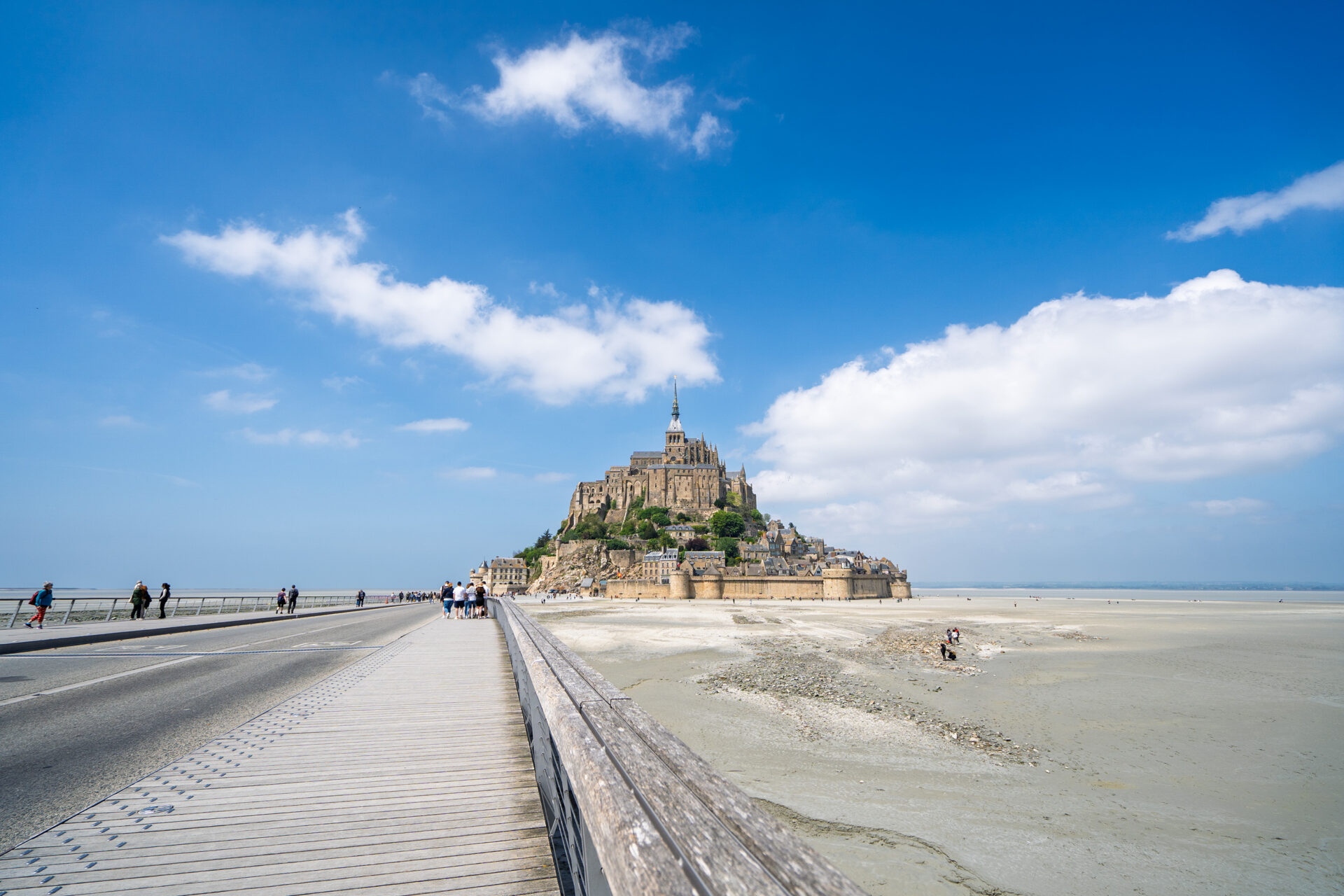 Mont Saint-Michel centered view with people walking on the Passerelle bridge on the left and mudflats with groups of people on the right 몽셍미셸을 중심으로, 왼쪽에는 파세렐 다리를 걷는 사람들, 오른쪽에는 드러난 뻘 위에 있는 사람들