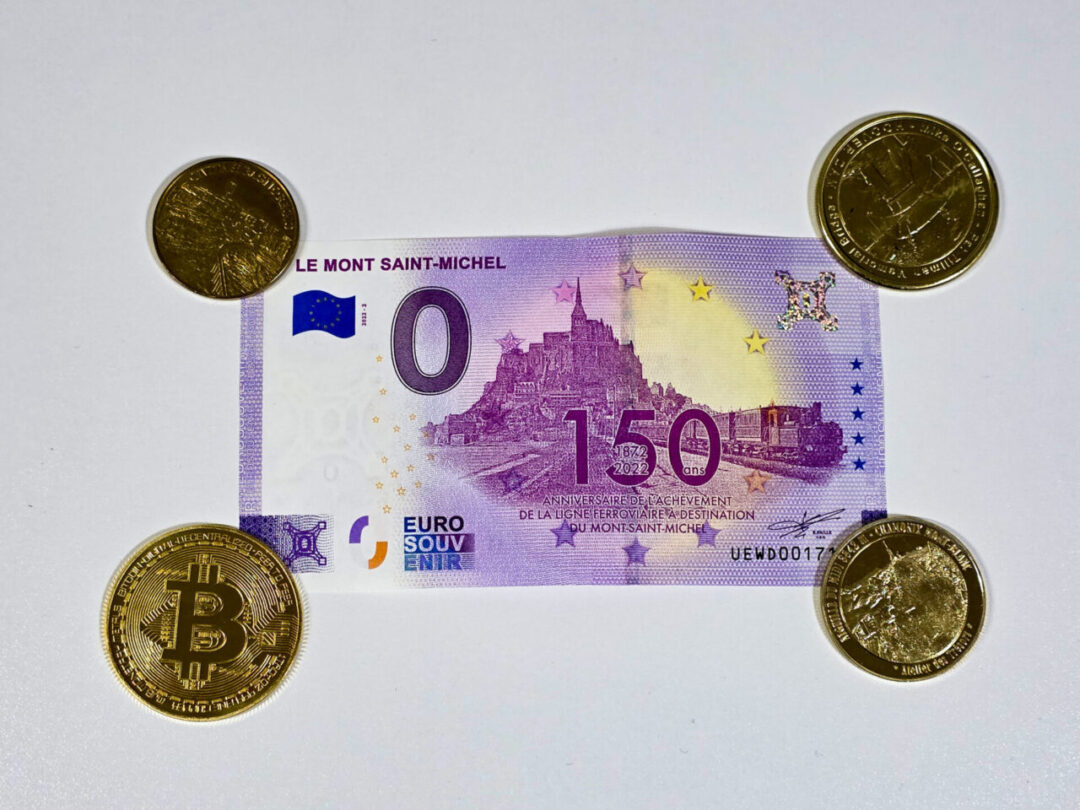 A Mont Saint-Michel souvenir 0-euro banknote, purchased as a keepsake, surrounded by various coins. 몽생미셸 기념품으로 구매한 0유로 지폐, 여러 동전과 함께 놓여 있는 모습.