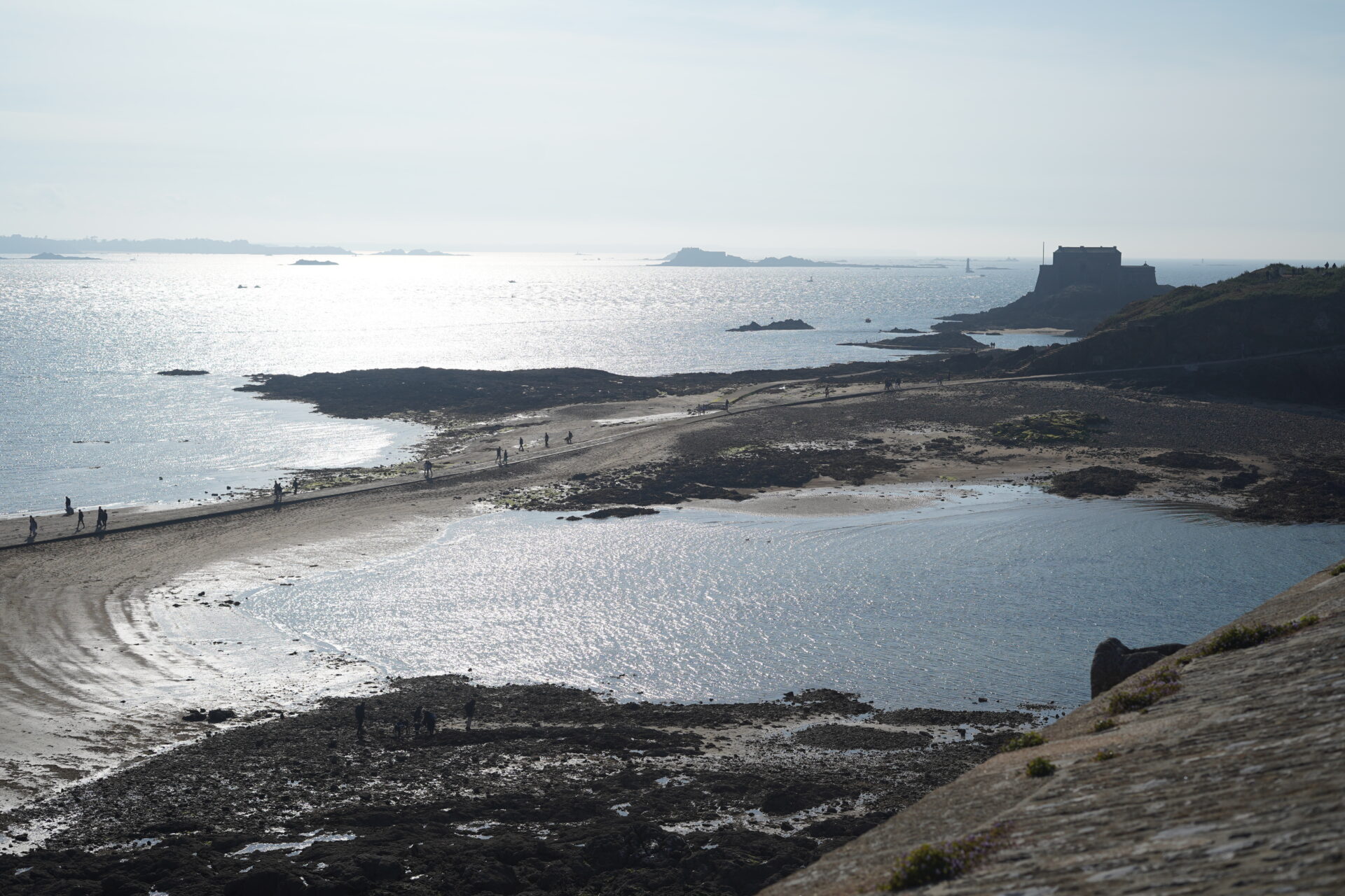 A pathway to Grand Bé in Saint-Malo, surrounded by water on both sides, with people walking towards the island. 생말로의 Grand Bé로 가는 길, 양쪽이 바닷물로 둘러싸이고 사람들이 섬을 향해 걸어가는 모습.