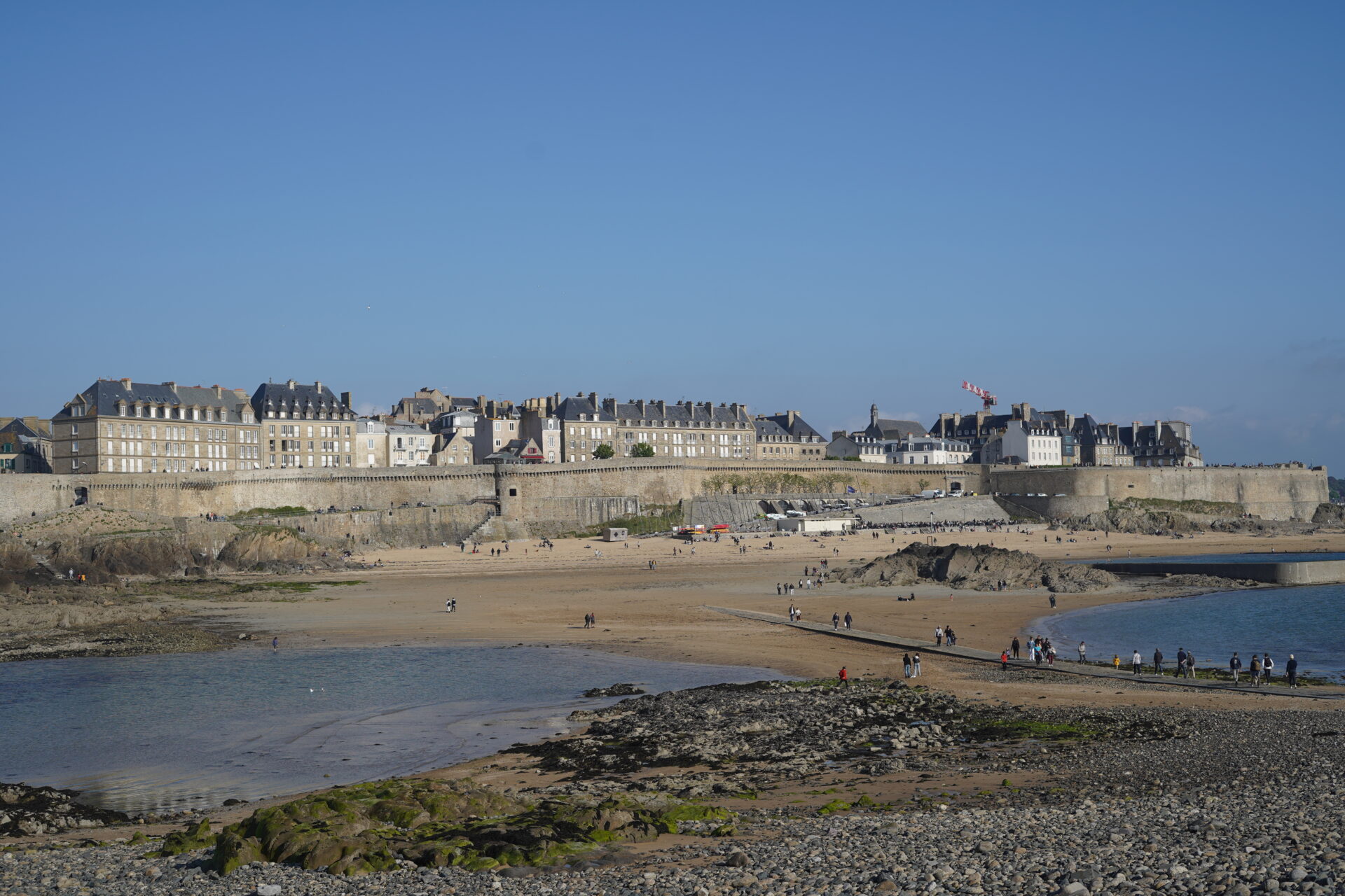 A view from Grand Bé island towards Saint-Malo, showing the tidal pathway filled with people, the Plage de Bon-Secours beach, and the city walls. Grand Bé 섬에서 생말로를 바라본 풍경으로, 사람들로 가득 찬 길과 Plage de Bon-Secours 해변, 그리고 도시 외벽이 펼쳐진 모습.