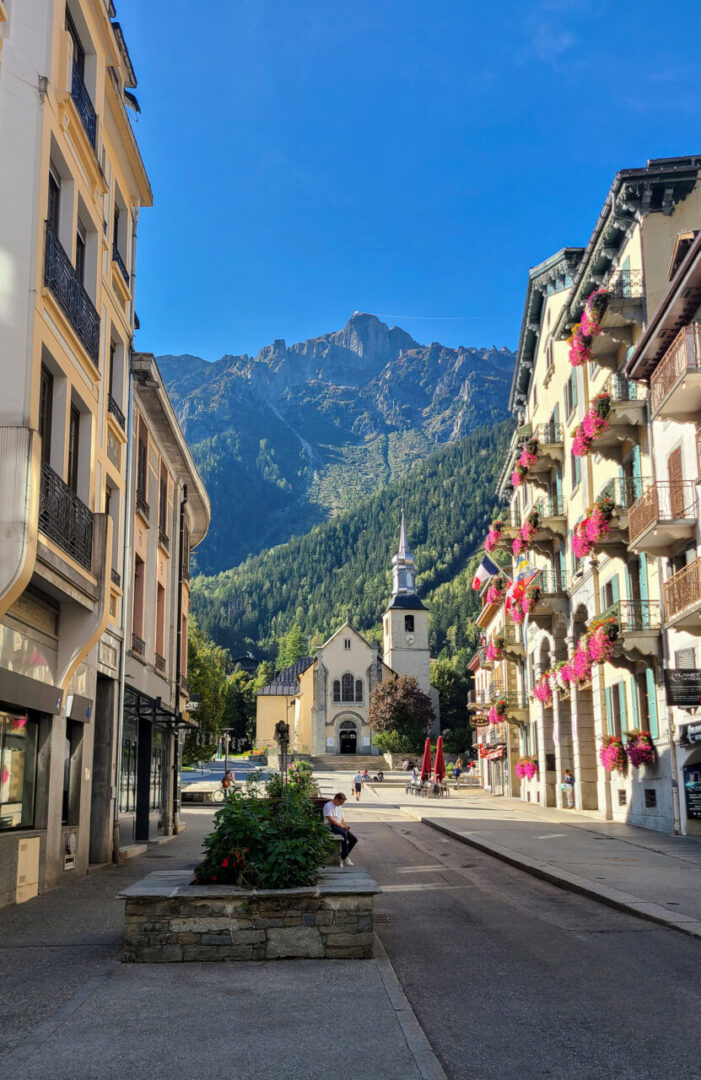 Église Saint-Michel in Chamonix with the Aiguilles Rouges mountain range in the background, featuring rugged peaks and a visible cable car line. 샤모니의 생 미셸 교회와 그 뒤로 펼쳐진 에귀유 루즈 산맥, 거친 봉우리들과 케이블카 라인이 보이는 풍경.