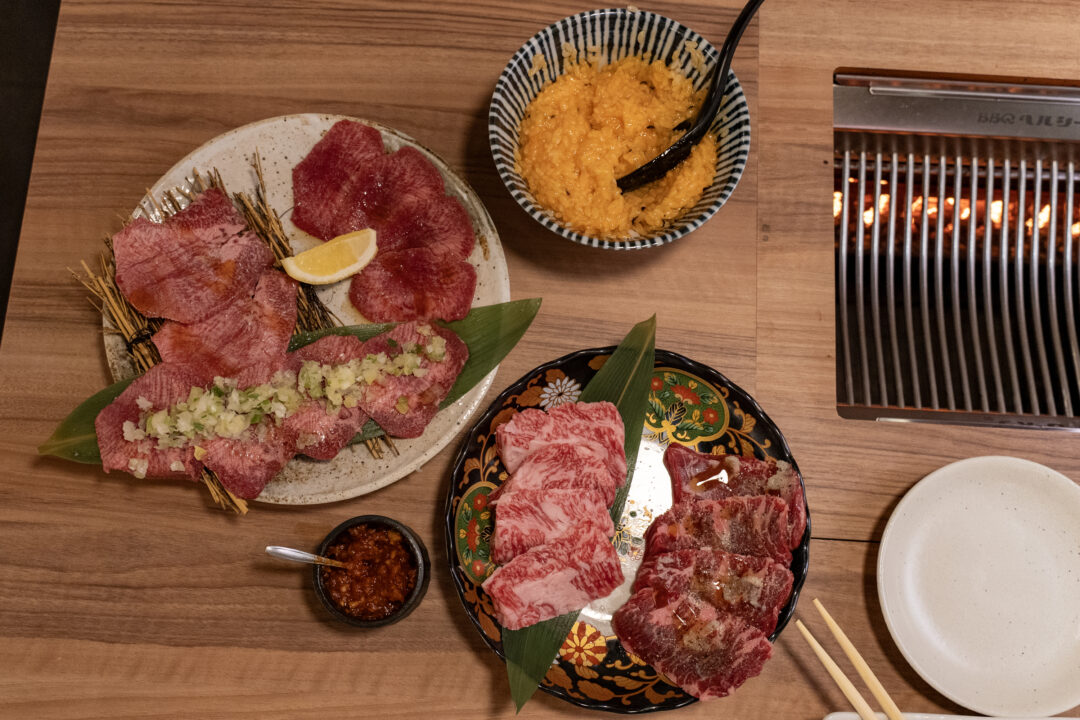 Full table setting at Yakiniku Inyen showing various cuts of beef including tongue and harami, egg rice, garlic miso and the smokeless grill. 신사이바시야키니쿠 인연의 테이블 세팅으로 우설과 하라미 등 다양한 소고기 부위, 계란밥, 갈릭 미소, 무연 그릴이 보임.