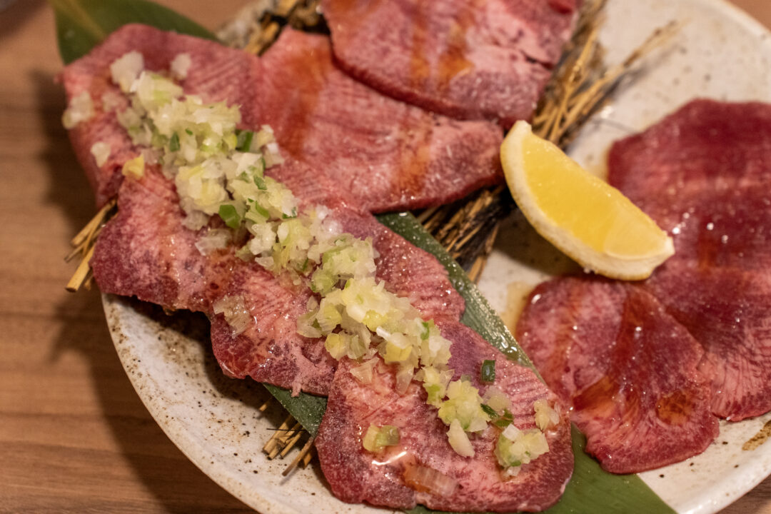 Beef tongue slices topped with chopped green onions and condiments on bamboo leaf at Yakiniku Inyen. 신사이바시야키니쿠 인연의 대나무 잎 위에 놓인 다진 파와 양념을 올린 우설 슬라이스.