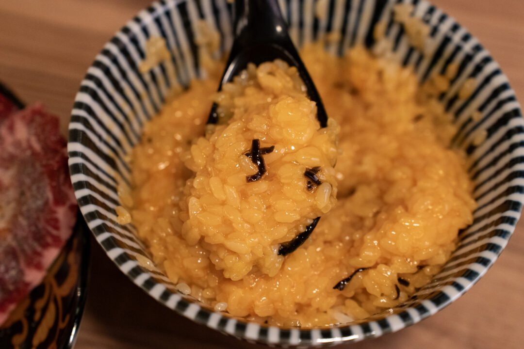 Close-up of fully mixed golden yellow TKG egg rice with seaweed flakes in a striped bowl at Yakiniku Inyen. 신사이바시야키니쿠 인연의 줄무늬 그릇에 담긴 완전히 비벼진 황금빛 TKG 계란밥과 다시마 조각.